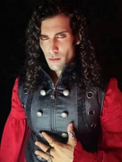 A man with long, curly black hair, wearing a black leather vest over a red blouse.