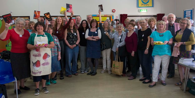 A large group of people gathered in a classroom to take a picture. Some hold up books and others hold plates of baked goods.